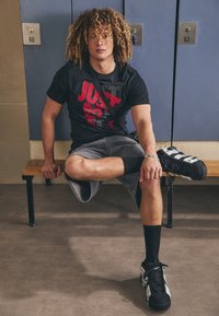 Svart kortermet t-skjorte med rød og grå tekst, grå shorts og svarte joggesko. Sitter på en trebenk ved blå skap.
