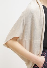 Beżowy szal o otwartym splotcie z luźnym krojem, miękką teksturą i szerokimi rękawami, opadający na czarną bluzkę, z widocznym detalem kieszeni.