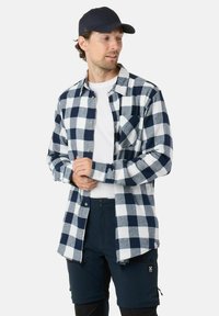 Marineblauw en witte geruite flanellen shirt met knopen, gedragen over een wit t-shirt, gecombineerd met donkere shorts. Een pet voegt een casual element toe.