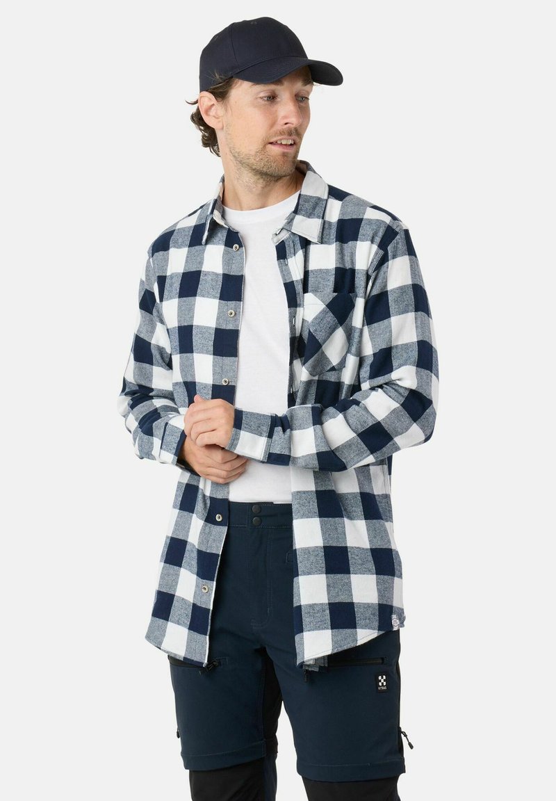 Marineblauw en witte geruite flanellen shirt met knopen, gedragen over een wit t-shirt, gecombineerd met donkere shorts. Een pet voegt een casual element toe.