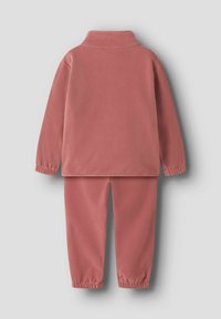 Survêtement en polaire de couleur rose pâle. Comprend un col haut, des poignets élastiques et une taille élastique. Texture lisse avec un design uni.