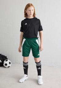 Must have musta särk mustade aktsentidega, rohelised lühikesed püksid, mustad jalgpallsokid hallide triipudega ja valged spikesid üksikasjalikus seisakus. Jalgpall läheduses.