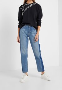 Sudadera negra con texto de logo diagonal en blanco combinada con jeans rectos de un tono azul claro que tienen paneles laterales más oscuros y zapatillas blancas.