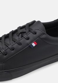 Svarta lädersneakers med platta svarta snörningar, slät yta, och en liten röd, vit och blå logotagg på sidan.