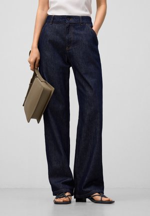 Mujer con jeans anchos de pierna oscura y sandalias negras con tiras, llevando un bolso taupe, sobre un fondo claro y liso.