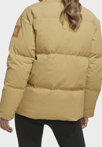 Žlutá péřová bunda s prošívaným designem, oversized střihem a logem na rukávu. Materiál vypadá hladce a má jemnou texturu.