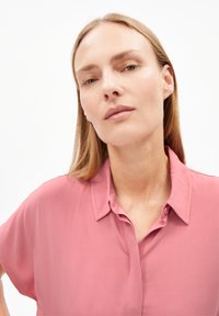 Helle rosa Bluse mit kurzen Ärmeln und Kragen-Design, glatter Textur und subtiler glänzender Optik, mit Knopfdetails entlang der Vorderseite.