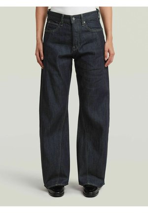 Person, die dunkelblaue Jeans mit weitem Bein, vorderen Taschen und schwarzen Schuhen trägt, stehend vor einem schlichten hellen Hintergrund.