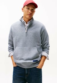 Maglione grigio in pile con zip a un quarto, tasca frontale e etichetta blu; abbinato a jeans scuri in denim e un cappellino rosso. La texture appare morbida.