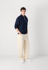 Felpa con zip color navy e colletto, indossata sopra pantaloni beige a gamba larga e sneaker bianche, con un piccolo dettaglio ricamato sul petto.