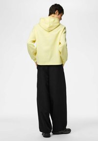 Sweat-shirt jaune à capuche avec une coupe décontractée, associé à un pantalon noir ample et des chaussures noires. Le tissu semble doux et texturé.