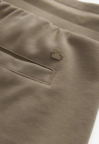 Jogging en tissu beige avec une ceinture texturée, une petite poche et un accent en métal à proximité de la poche. Matière lisse et extensible.