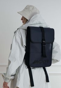 Zaino impermeabile blu marina con chiusura a patta, spallacci regolabili e logo della marca testurizzato sul fronte. Indossato sopra un cappotto bianco.