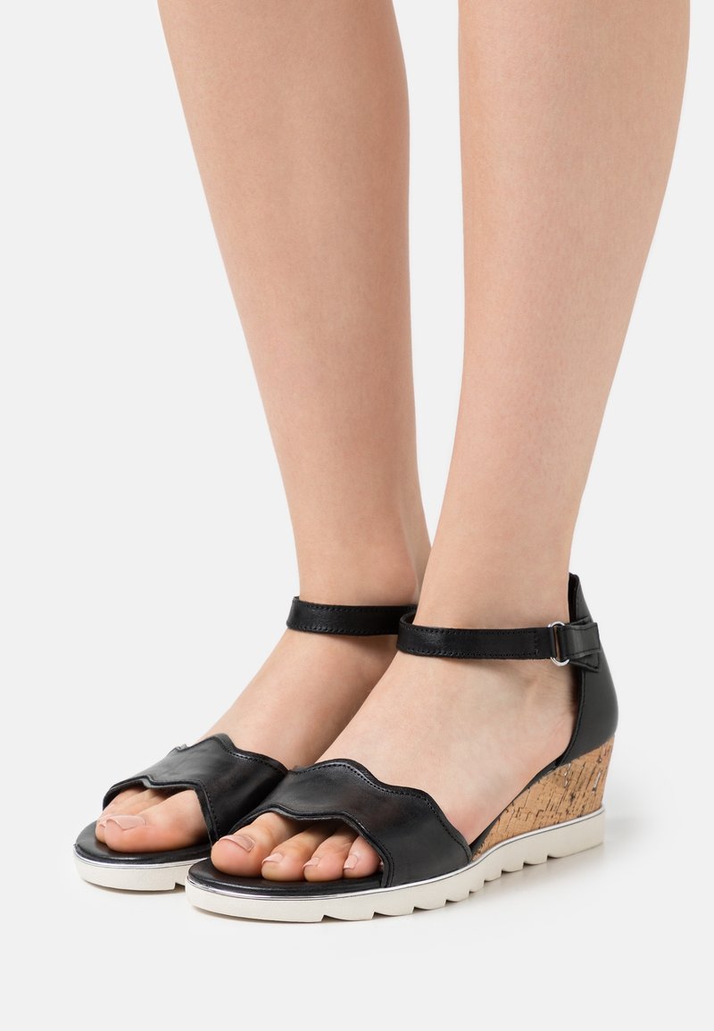 Des pieds portant des sandales en cuir noir à bout ouvert avec des brides à la cheville et des talons compensés en liège sur une surface blanche.