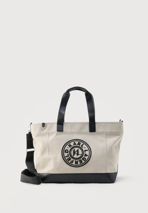 Béžová taška přes rameno s černými úchyty a popruhem, černým lemováním na spodní části a kulatým logem "Karl Lagerfeld" uprostřed přední strany.