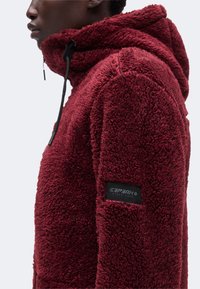 Profil latéral d'une personne portant une veste polaire à capuche texturée rouge foncé avec un patch logo Icepeak noir sur la manche.