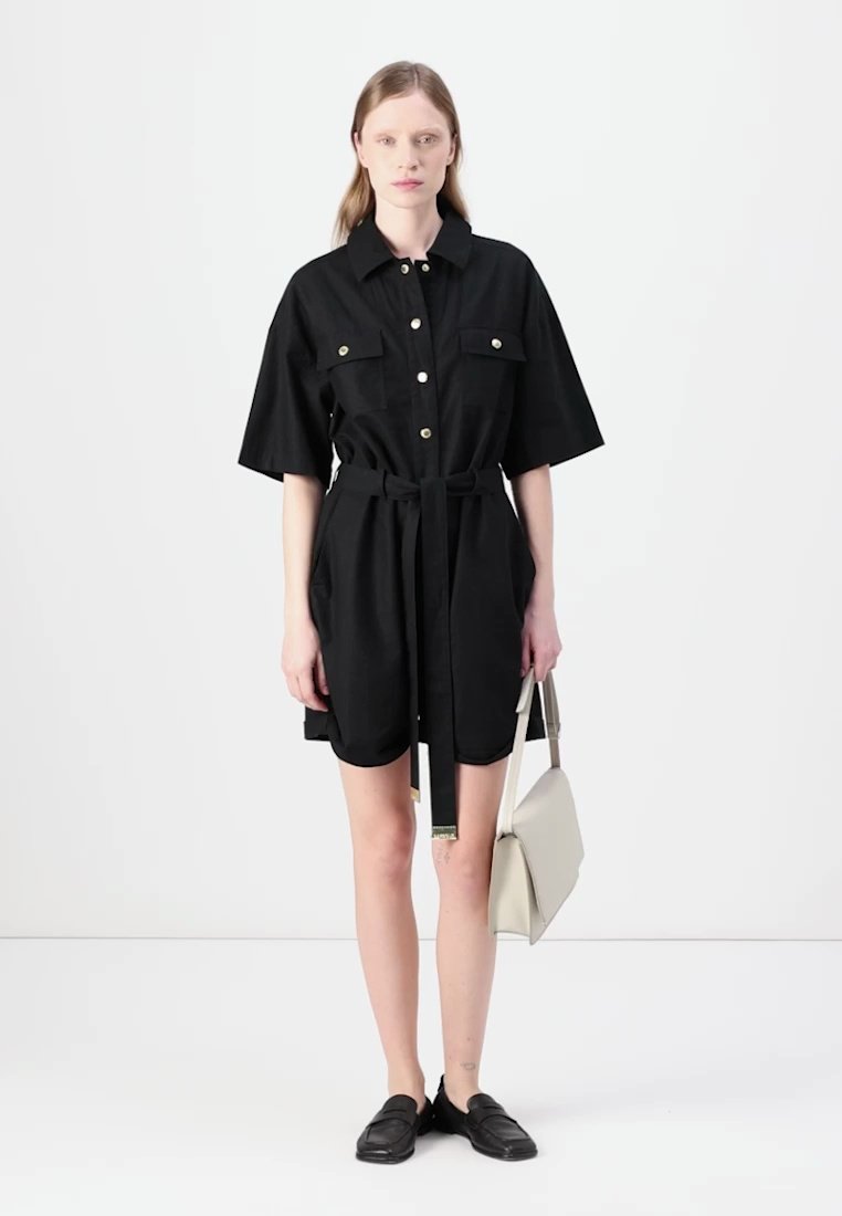 Robe noire à manches courtes avec un col boutonné, deux poches poitrine, une ceinture à la taille et un tissu lisse. Assortie à un sac à épaule de couleur claire.