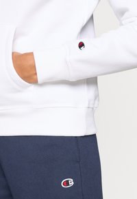 Λευκό φούτερ με μακριά μανίκια και ριμπ μανσέτες, με λογότυπο στο μανίκι. Συνδυάζεται με σκούρο μπλε παντελόνι που έχει λογότυπο.