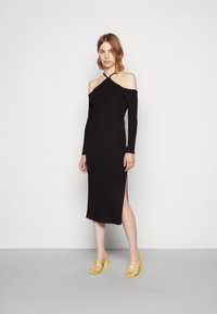 Vestido preto com ombros à mostra, mangas compridas, design justo, comprimento até à meia panturrilha, detalhe de fenda lateral. Usado com saltos amarelos com tiras.