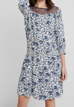 Kobieta w białej sukience z niebieskim wzorem kwiatowym, przezroczystym, czarnym, kropkowanym dekoltem, rękawami za łokieć i marszczoną elastyczną talią.