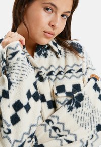 Fleece overhemdjack met een crème basis en marineblauwe patronen, voorzien van zwarte knopen, een borstzak en een leren label op de mouw.