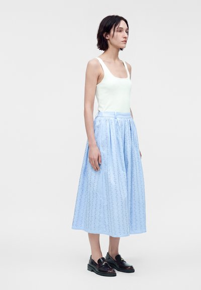 Femme portant un haut blanc sans manches, une jupe midi bleu clair avec motif perforé et des mocassins noirs, debout devant un fond uni.