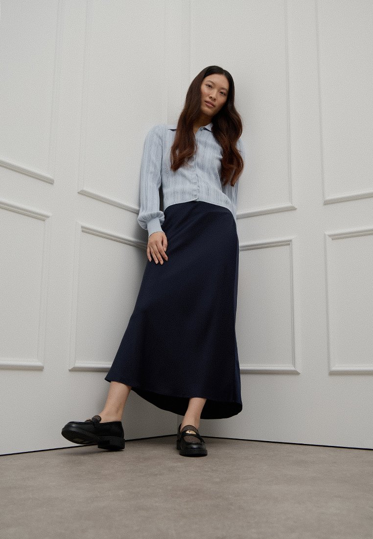 Vrouw met lang haar leunt tegen witte paneelwanden, gekleed in een lichtblauwe blouse, een marineblauwe midi-rok en zwarte instappers.