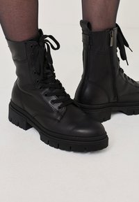 Black leather ankle boots featuring a lace-up front, side zipper, and chunky outsole with textured tread pattern. Textured details on the upper.