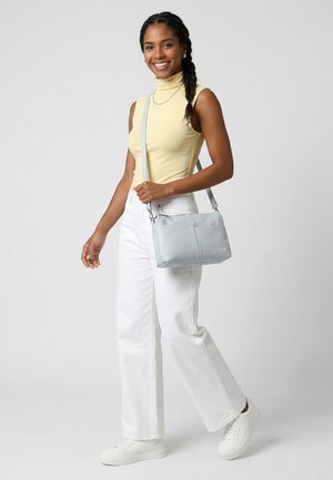 Donna sorridente con capelli intrecciati, indossa un dolcevita giallo senza maniche, pantaloni bianchi, scarpe da ginnastica bianche e porta una borsa a tracolla grigio chiaro.