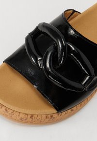 Sandale à glissière en vernis noir avec un accent de chaîne chunky. La semelle en liège texturé a une teinte marron clair, contrastant avec la partie supérieure noire.