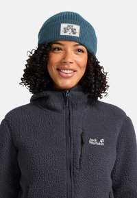 Vrouw met een blauwe gebreide beanie en een donkergrijze fleece jack met rits, voorzien van Jack Wolfskin-markering, die glimlacht en iets naar rechts kijkt.