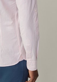 Camicia bianca con sottili righe rosa, caratterizzata da un orlo posteriore arrotondato e polsini con bottoni. Tessuto liscio e vestibilità su misura.