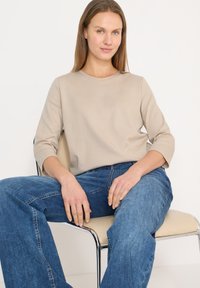 Chemise beige à manches longues avec un col rond, associée à un jean en denim bleu à jambes larges. Assis sur une chaise chromée avec une tapisserie neutre.