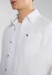Camicia bianca a maniche lunghe realizzata in tessuto leggero, con una tasca sul petto decorata da un piccolo emblemma a forma di bandiera, bottoni argentati e una texture morbida.