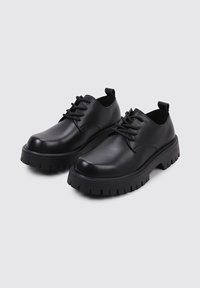 Zapatos de cuero negro con cordones, punta redondeada, suela gruesa y estriada, y un diseño minimalista. Presentan una tira en el talón para facilitar su puesta.