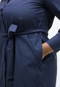 Χέρι γυναίκας στην τσέπη φορέματος σε ναυτικό μπλε χρώμα με ζώνη, κουμπιά, μακριά μανίκια και γιακά.