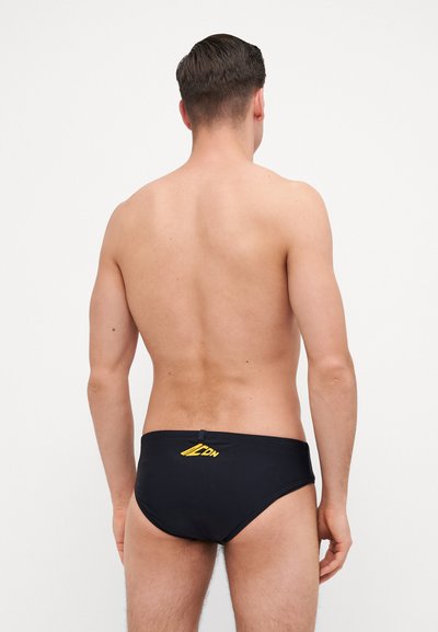 Caleçons de bain noirs avec un logo jaune et une taille élastique, montrant la vue arrière d'une silhouette masculine sans autre vêtement.