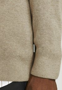 Maglione khaki con un motivo a maglia testurizzato. Presenta un polsino a coste e un piccolo etichetta del marchio sul lato. Materiale morbido e caldo senza loghi visibili.