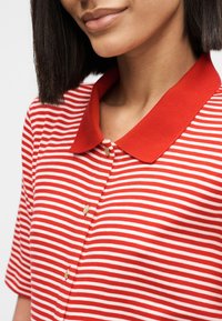 Γυναίκα με κοντά σκούρα μαλλιά που φοράει κόκκινο και λευκό ριγέ πόλο πουκάμισο με χρυσά κουμπιά, εμφανιζόμενη από τον λαιμό έως το μέσο του κορμού.