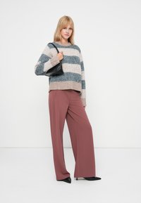 Blonde sieviete, tērpusies platām mauve krāsas biksēm, svītrainā smilškrāsas un zilā džemperī, melnās norādītās apavās, tur mazu melnu somiņu uz pleca.