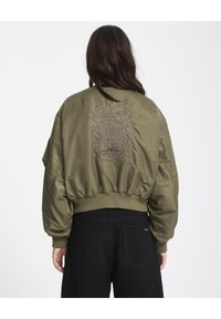 Olivengrøn bomberjakke med oversized ærmer, ribbede manchetter og broderet design på ryggen. Båret med sorte bukser.
