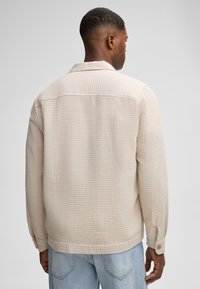 Camicia beige a maniche lunghe con texture e motivo a griglia, dotata di colletto e polsini con bottoni, abbinata a jeans in denim blu chiaro.