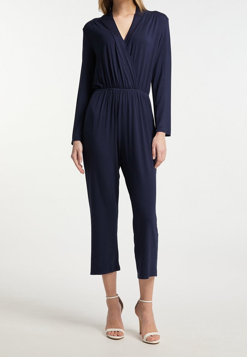 Femme portant une combinaison bleu marine à manches longues avec un devant croisé et des jambes raccourcies, associée à des sandales à talons hauts beige clair.
