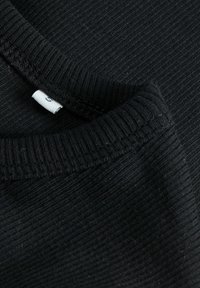 Schwarzer gerippter Stoff, der eine Nahaufnahme des Kragens und des Etiketts eines Kleidungsstücks zeigt. Die Textur ist glatt mit sichtbaren Nähdetails.