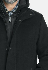 Abrigo negro de mezcla de lana con una capa interior con cremallera, botones a presión y capucha. Presenta una textura acolchada y un brillo sutil.