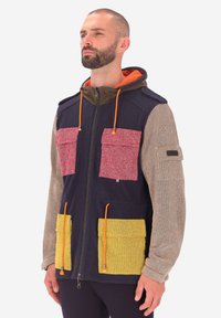 Uomo che indossa un giubbotto con cappuccio a blocchi di colore, con pannelli blu navy, beige, rosso e giallo, lacci arancioni e molteplici tasche anteriori.