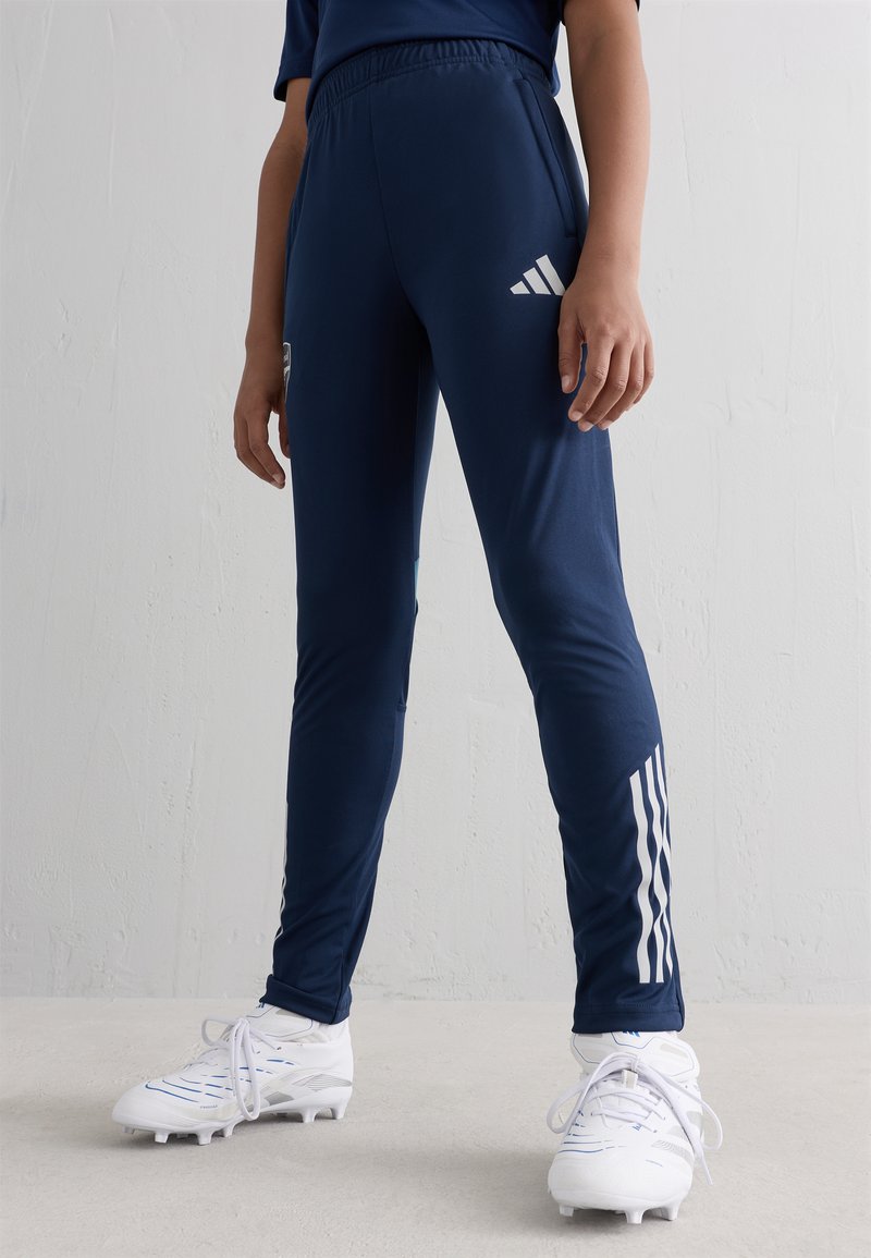 Persona, tērpta tumši zilos Adidas treniņbiksēs un baltos sporta apavos, stāv pret tukšu gaiši pelēku strukturētu sienu.