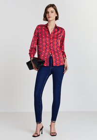 Frau in einer roten Bluse mit floralem Muster, dunkelblauen Skinny-Jeans, schwarzen High-Heelsandalen und hält eine schwarze Clutch-Tasche.