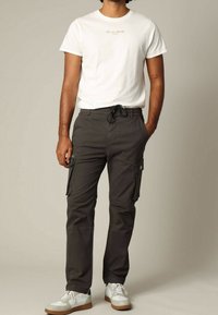 Biała koszulka z krótkim rękawem z napisem, zestawiona z ciemnymi spodniami typu cargo z bocznymi kieszeniami. Model ma na sobie jasne buty z brązowymi elementami.