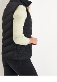 Černá prošívaná vesta s vysokým límcem, nošená přes světle zelený přízevkový svetr. Obsahuje diagonální švy a boční kapsy.